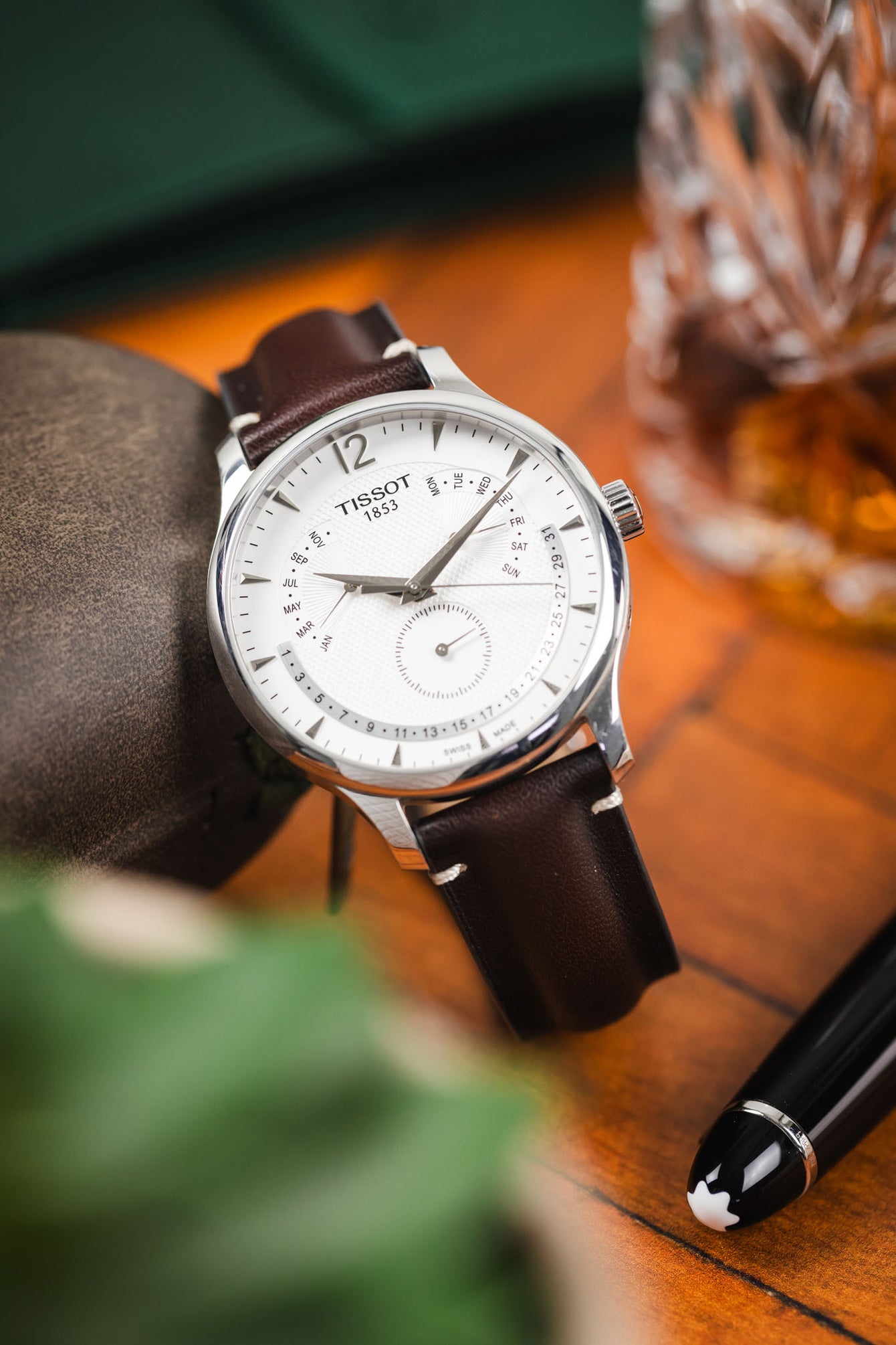 Close-up of a Tissot watch with a white dial and brown leather strap, styled on a wooden surface beside a whisky glass and pen.