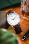 Close-up of a Tissot watch with a white dial and brown leather strap, styled on a wooden surface beside a whisky glass and pen.