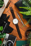 Flat-lay of a Tissot watch with a white dial and brown leather strap, arranged with a camera, tools, pen, and greenery on a wooden surface.