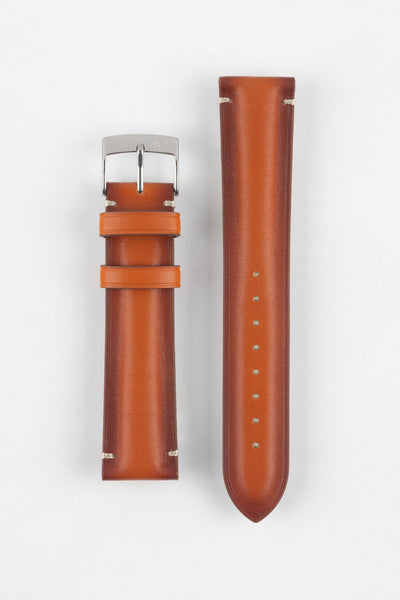 Two warm brown leather watch strap pieces displayed vertically on a light grey background. One strap section features a polished silver tang buckle with an engraved logo and two matching leather keepers, while the other shows a tapered end with evenly spaced adjustment holes. The leather has a smooth, slightly burnished finish with subtle tonal variation and light contrast stitching near the lugs, highlighting its craftsmanship.