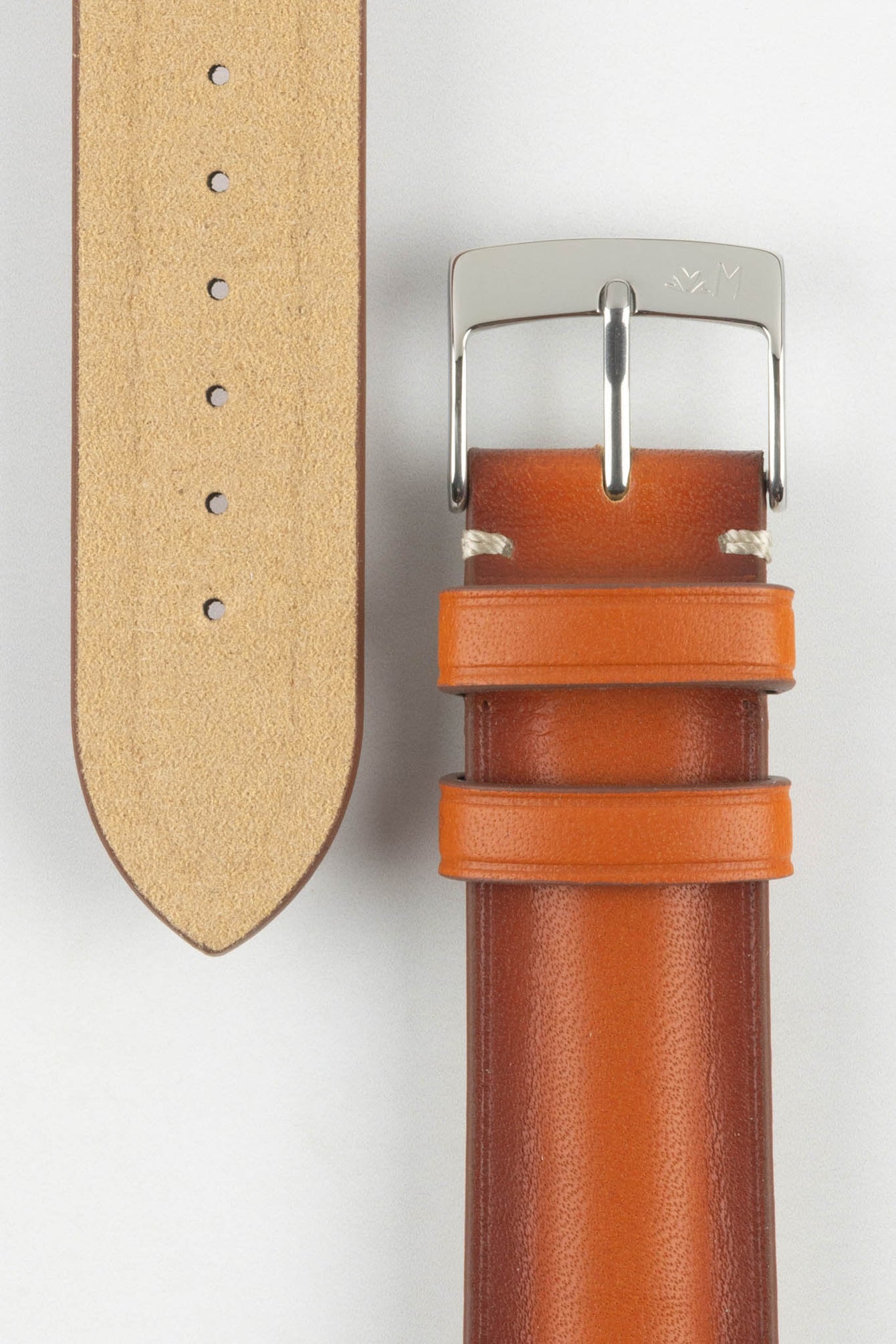 Close-up of a warm brown leather watch strap on a light grey background, showing one section with a polished silver tang buckle engraved with a small logo and two matching leather keepers. Beside it, the opposite strap section reveals the tan suede underside with a vertical row of evenly spaced adjustment holes. The leather has a smooth, lightly burnished finish with subtle tonal variation and light contrast stitching near the buckle, emphasizing the strap’s craftsmanship.