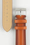 Close-up of a warm brown leather watch strap on a light grey background, showing one section with a polished silver tang buckle engraved with a small logo and two matching leather keepers. Beside it, the opposite strap section reveals the tan suede underside with a vertical row of evenly spaced adjustment holes. The leather has a smooth, lightly burnished finish with subtle tonal variation and light contrast stitching near the buckle, emphasizing the strap’s craftsmanship.