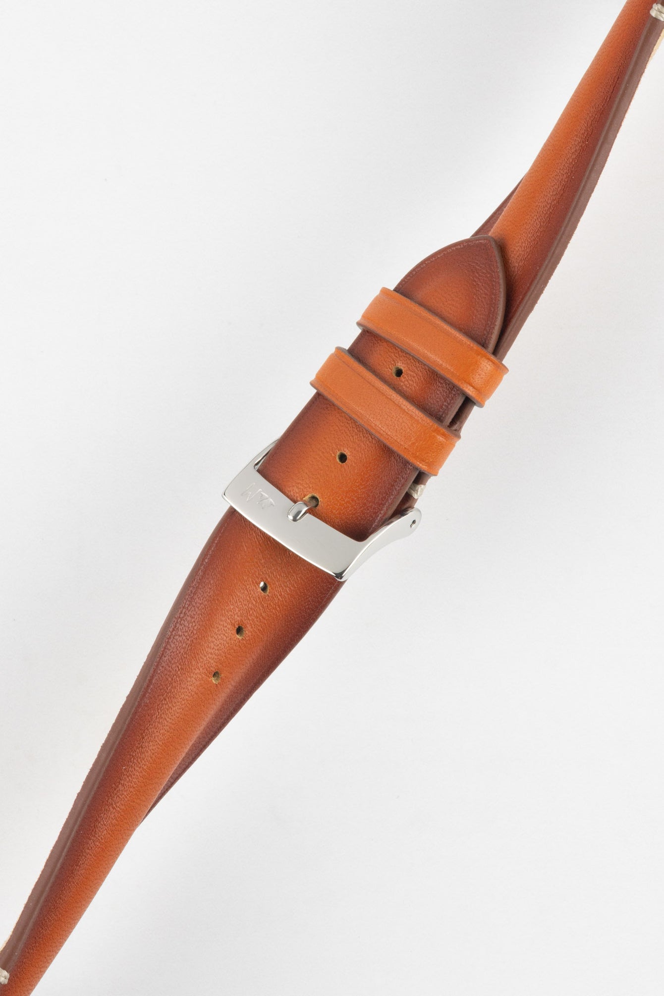 Angled close-up of a warm brown leather watch strap laid on a light grey background, featuring a polished silver tang buckle with an engraved logo and two matching leather keepers. The strap shows a smooth, slightly burnished finish with visible tonal variation, along with a row of circular adjustment holes along its length. Subtle contrast stitching near the lugs and light creasing in the leather highlight the strap’s texture and craftsmanship.