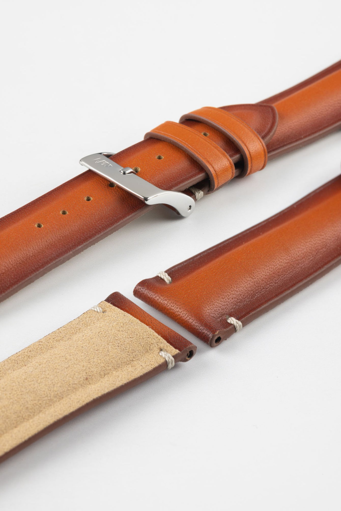Angled close-up of a warm brown leather watch strap displayed on a light grey background, with multiple strap sections arranged side by side. One section features a polished silver tang buckle with an engraved logo and two matching leather keepers, while the others show the smooth, slightly burnished leather exterior and the tan suede underside near the lugs. The strap includes evenly spaced adjustment holes and subtle contrast stitching, highlighting the craftsmanship and construction.
