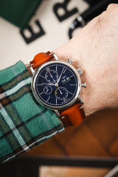 Close-up of an IWC chronograph with a black dial and brown leather strap, worn on a wrist, with a green plaid sleeve in view.