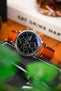 Close-up of an IWC chronograph watch with a black dial and brown leather strap, resting on books with a blurred whisky glass and greenery in the background.