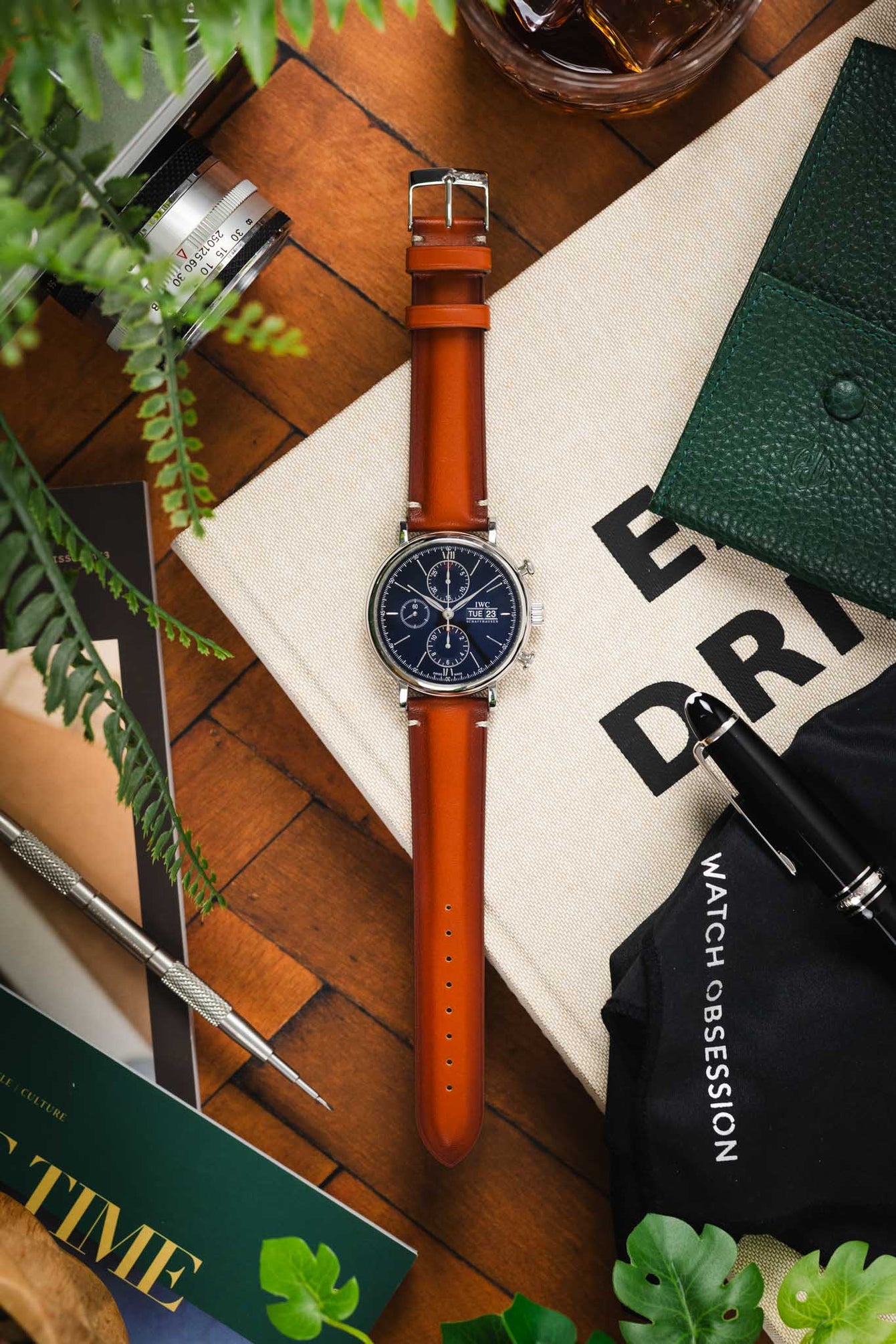 Flat-lay of an IWC chronograph with a brown leather strap, surrounded by stationery, books, greenery, and a drink on a wooden surface.