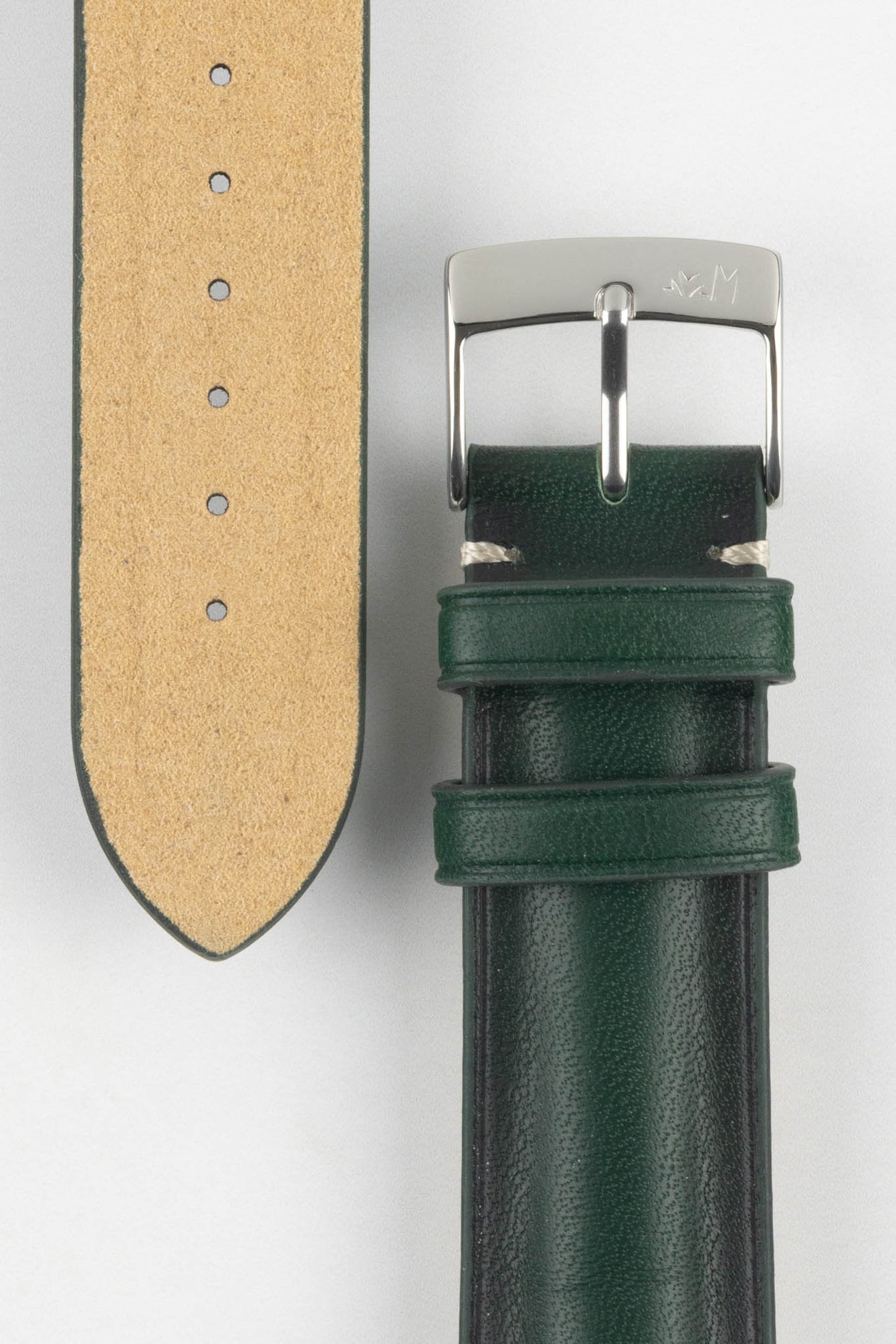 Close-up of a dark green leather watch strap on a light grey background, showing one section with a polished silver tang buckle featuring an engraved logo and two matching leather keepers. Beside it, the opposite strap section reveals the tan suede underside with a vertical row of evenly spaced adjustment holes. The leather has a smooth, slightly pebbled finish with light contrast stitching near the buckle, highlighting the strap’s construction.