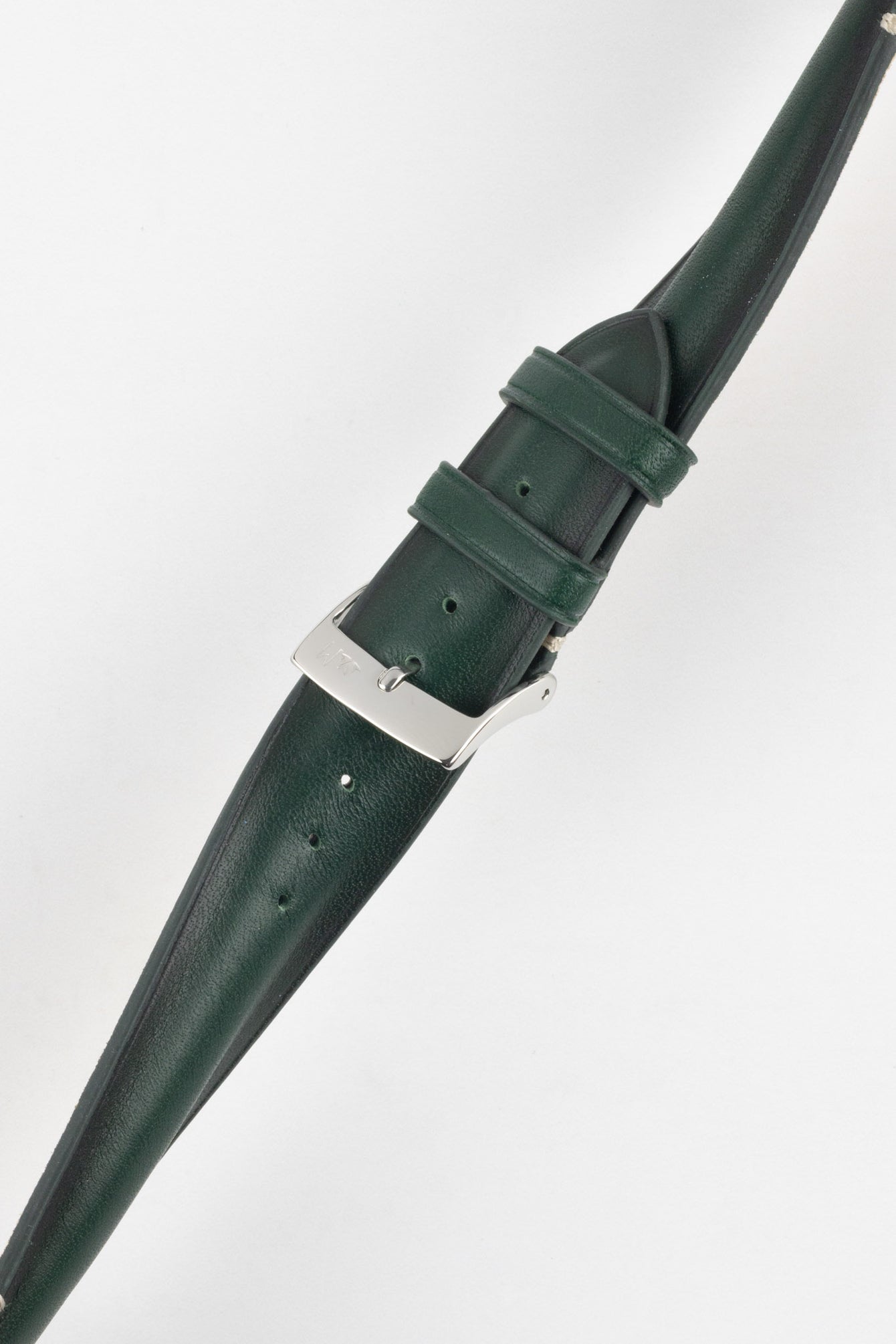 Angled close-up of a dark green leather watch strap set against a light grey background, featuring a polished silver tang buckle with an engraved logo and two matching leather keepers. The strap shows a smooth, slightly pebbled texture with a row of adjustment holes along its length, and subtle contrast stitching near the lugs, highlighting its refined construction and finish.