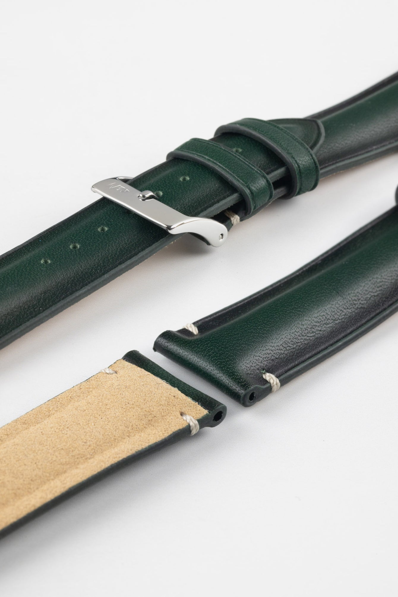 Angled close-up of a dark green leather watch strap displayed on a light grey background, with multiple strap sections arranged side by side. One section features a polished silver tang buckle with an engraved logo and two matching leather keepers, while the others showcase the smooth, slightly pebbled green leather exterior and the tan suede underside near the lugs. The strap includes evenly spaced adjustment holes and subtle contrast stitching, highlighting the craftsmanship and construction details.