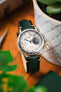 Studio Underd0g Bicompax watch with a textured silver dial and dark green leather strap, styled beside a plant pot on a wooden surface.