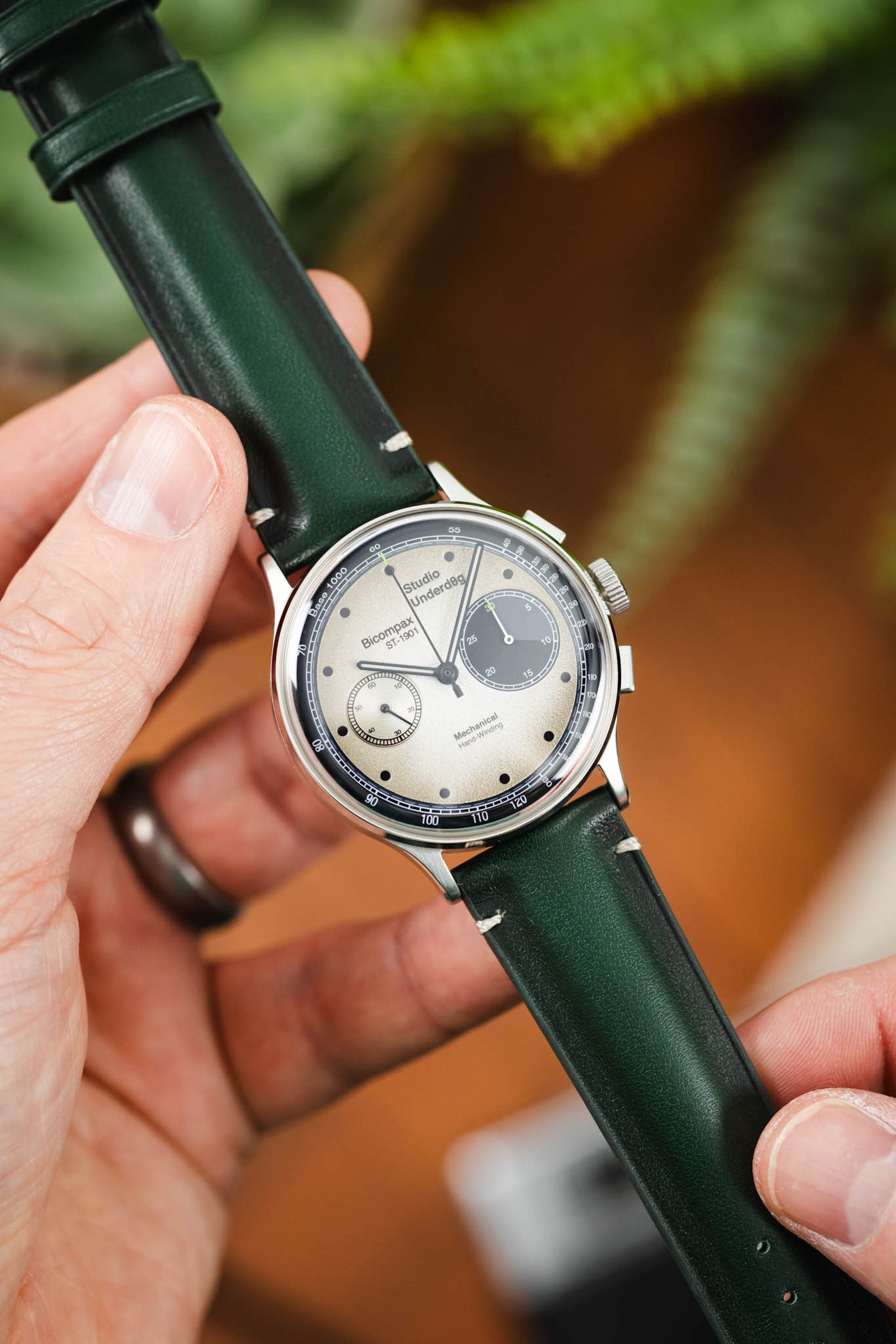 Close-up of a Studio Underd0g Bicompax chronograph watch with a textured silver dial, featuring two subdials, slim black markers, and tachymeter-style detailing around the edge. The polished steel case is paired with a dark green leather strap with white stitching. The watch is held between fingers, with the strap extended, while a softly blurred background of greenery and warm tones highlights the strap’s smooth finish and rich colour.