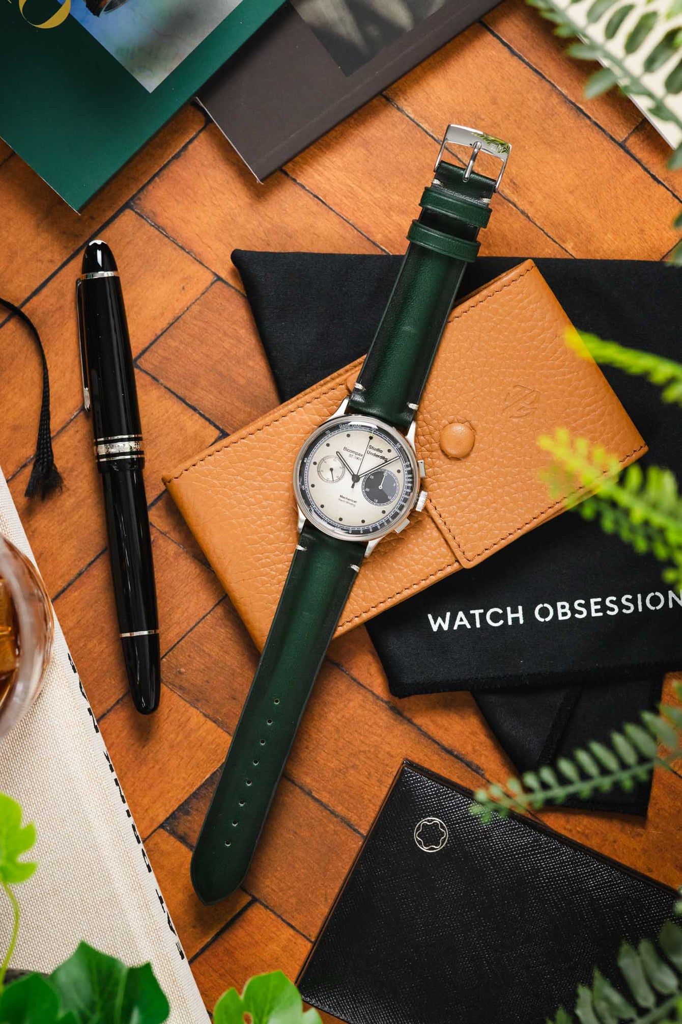 Flat-lay of a Studio Underd0g Bicompax watch with a dark green leather strap, placed on a tan leather case and surrounded by pens, notebooks, and greenery on a wooden surface.