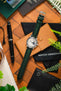 Flat-lay of a Studio Underd0g Bicompax watch with a dark green leather strap, placed on a tan leather case and surrounded by pens, notebooks, and greenery on a wooden surface.