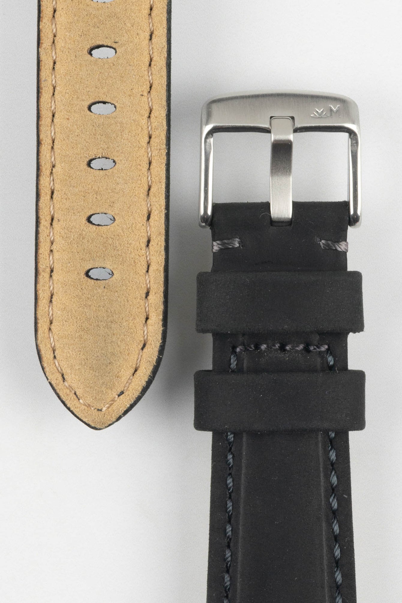Close-up of a black leather watch strap on a light grey background, showing one section with a brushed silver tang buckle featuring an engraved logo and two leather keepers. Beside it, the opposite strap section reveals the tan suede underside with visible stitching along the edges and a row of evenly spaced oval adjustment holes. The leather has a matte, slightly textured finish with dark contrast stitching, emphasizing durability and craftsmanship.
