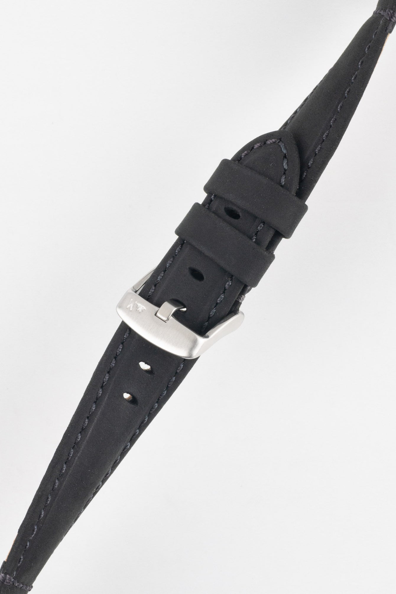 Angled close-up of a black leather watch strap on a light grey background, featuring a matte, slightly textured finish with dark grey stitching along the edges. The strap includes a brushed silver tang buckle with an engraved logo and two leather keepers. A series of evenly spaced oval adjustment holes and a tapered tip are visible, highlighting the strap’s structured design and durable construction.