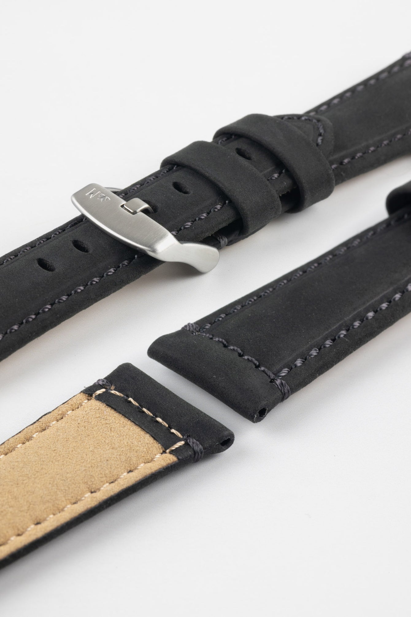 Angled close-up of a black leather watch strap displayed on a light grey background, with the strap separated into multiple sections. One section features a brushed silver tang buckle with an engraved logo and two leather keepers, while adjacent pieces show the matte, slightly textured black leather exterior with dark grey stitching and the tan suede underside near the lugs. Oval adjustment holes, clean stitching lines, and the strap’s tapered form highlight its durable construction and contemporary design.