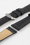 Angled close-up of a black leather watch strap displayed on a light grey background, with the strap separated into multiple sections. One section features a brushed silver tang buckle with an engraved logo and two leather keepers, while adjacent pieces show the matte, slightly textured black leather exterior with dark grey stitching and the tan suede underside near the lugs. Oval adjustment holes, clean stitching lines, and the strap’s tapered form highlight its durable construction and contemporary design.