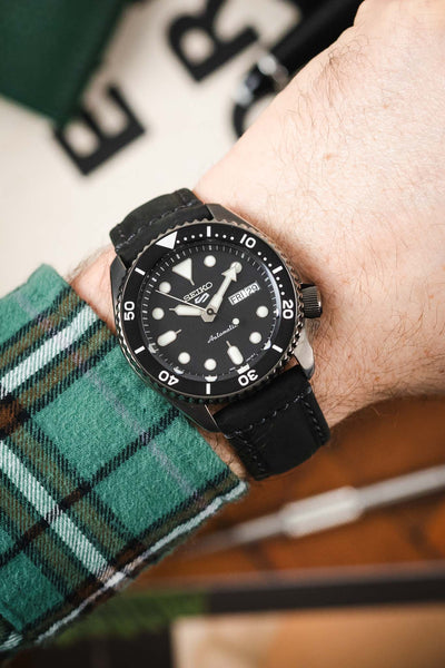 Close-up of a Seiko dive watch with a black dial and rotating bezel, worn on a wrist with a black leather strap and a green plaid sleeve.