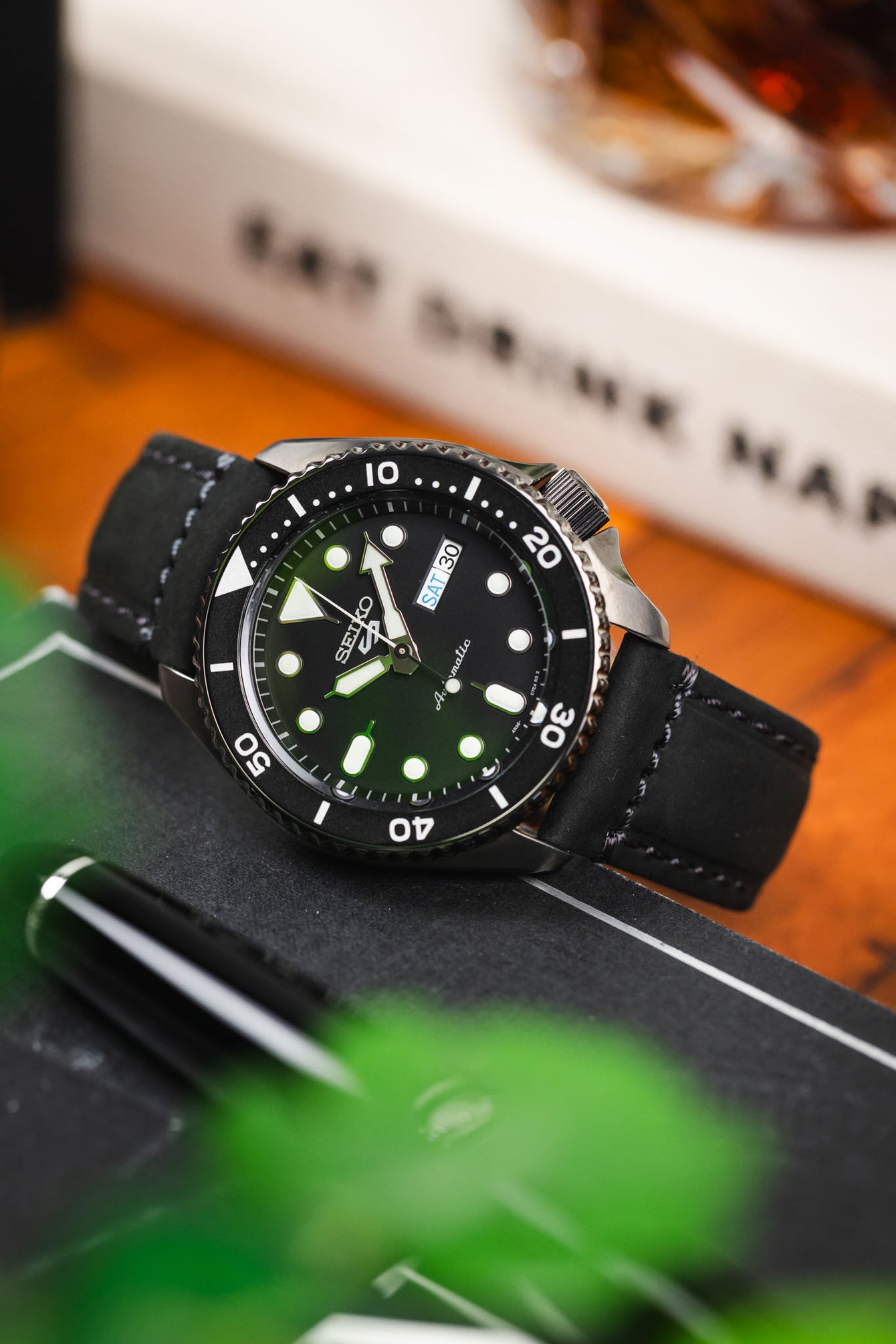 Close-up of a Seiko dive watch with a black dial and rotating bezel on a black leather strap, propped against a book with blurred greenery and warm-toned background elements.