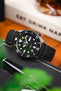 Close-up of a Seiko dive watch with a black dial and rotating bezel on a black leather strap, propped against a book with blurred greenery and warm-toned background elements.