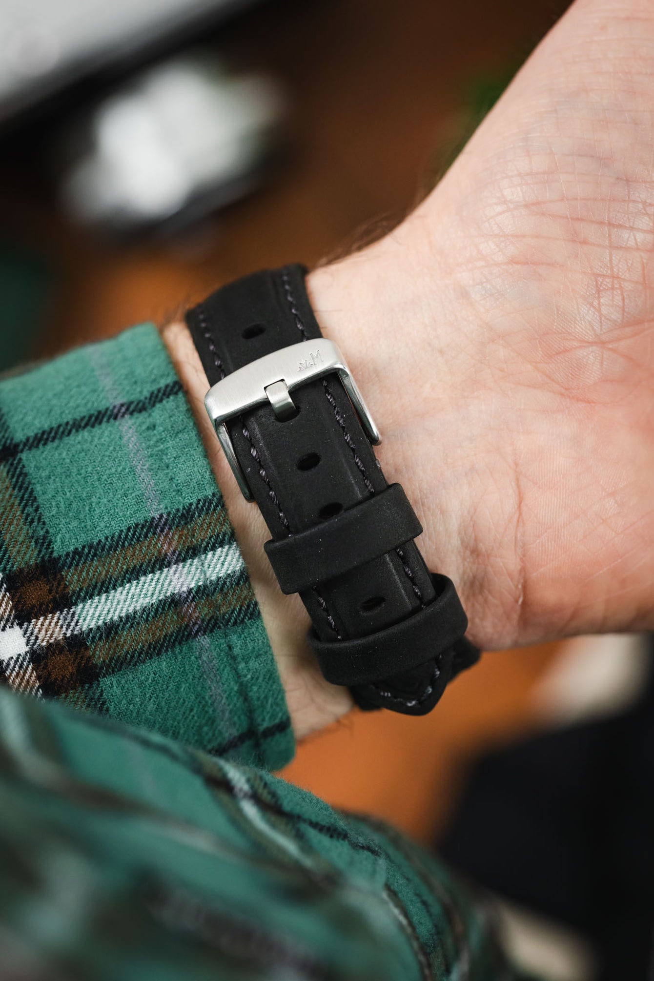 Close-up of a black leather watch strap with contrast stitching, secured on a wrist with a brushed silver buckle and keeper loops, worn with a green plaid sleeve.