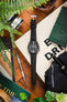 Flat-lay of a Seiko dive watch on a black leather strap, arranged with a camera, pen, tools, greenery, and accessories on a wooden surface.