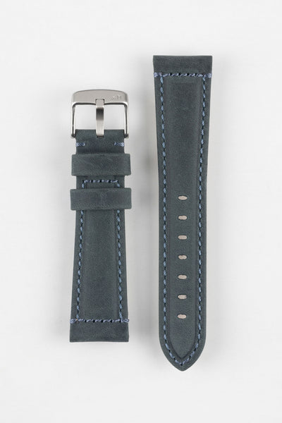Two blue-grey leather watch strap pieces displayed vertically on a light grey background, featuring a matte, finely textured finish with contrast light blue stitching along the edges. One strap section includes a brushed silver tang buckle with an engraved logo and two leather keepers, while the other shows a tapered end with evenly spaced oval adjustment holes. The clean stitching and structured shape highlight the strap’s modern, durable design.