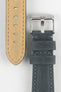 Close-up of a blue-grey leather watch strap on a light grey background, showing one section with a brushed silver tang buckle featuring an engraved logo and two leather keepers. To the left, the opposite strap section reveals the tan suede underside with visible stitching along the edges and a row of evenly spaced oval adjustment holes. The outer leather has a matte, finely textured finish with contrast light blue stitching, emphasizing the strap’s construction and modern design.