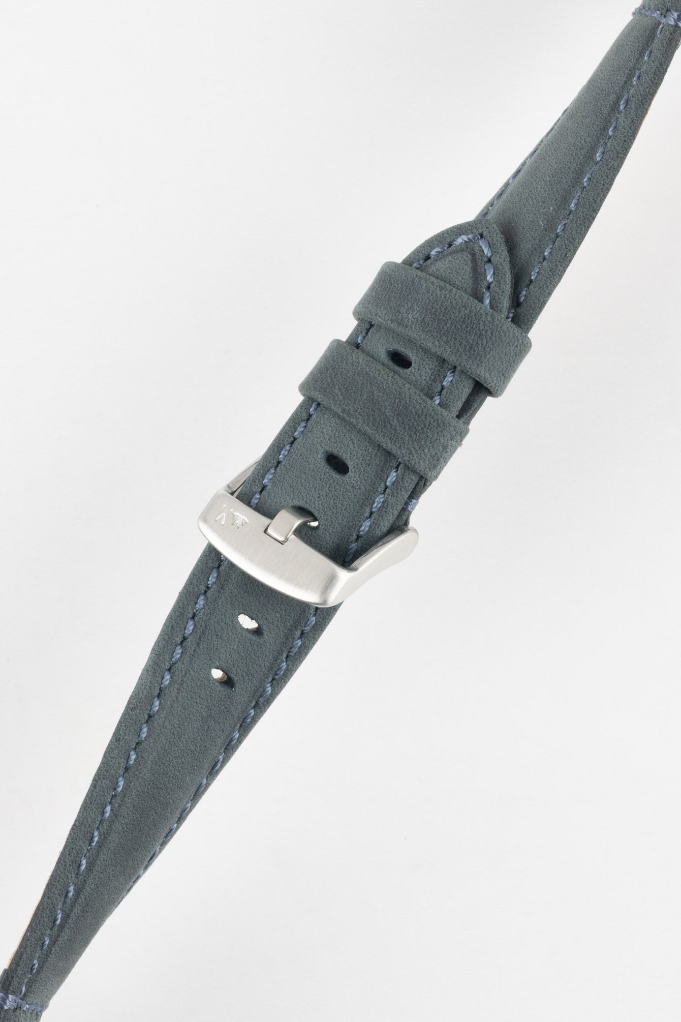 Close-up of a blue-grey leather watch strap on a light grey background, showing one section with a brushed silver tang buckle featuring an engraved logo and two leather keepers. To the left, the opposite strap section reveals the tan suede underside with visible stitching along the edges and a row of evenly spaced oval adjustment holes. The outer leather has a matte, finely textured finish with contrast light blue stitching, emphasizing the strap’s construction and modern design.