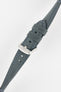 Close-up of a blue-grey leather watch strap on a light grey background, showing one section with a brushed silver tang buckle featuring an engraved logo and two leather keepers. To the left, the opposite strap section reveals the tan suede underside with visible stitching along the edges and a row of evenly spaced oval adjustment holes. The outer leather has a matte, finely textured finish with contrast light blue stitching, emphasizing the strap’s construction and modern design.