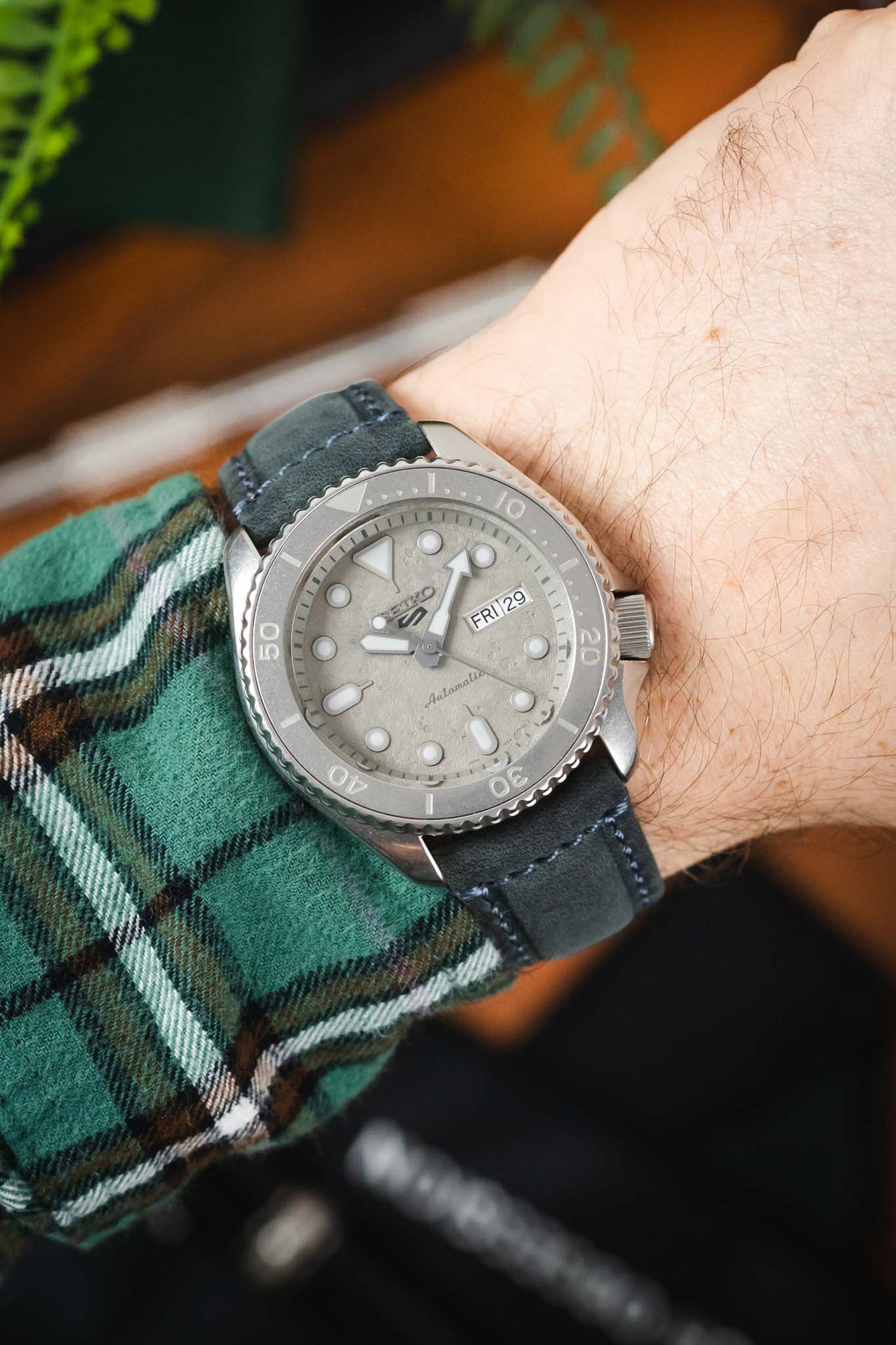 Close-up of a Seiko-style dive watch with a textured light dial and rotating bezel, worn on a wrist with a blue leather strap and a green plaid sleeve visible.