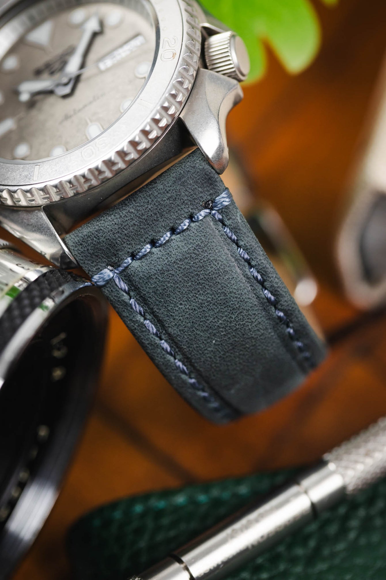 Macro close-up of a blue leather watch strap with contrasting light stitching attached to a brushed steel dive watch case, highlighting the textured grain, stitching detail, and bezel edge, with blurred tools and greenery in the background.