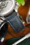 Macro close-up of a blue leather watch strap with contrasting light stitching attached to a brushed steel dive watch case, highlighting the textured grain, stitching detail, and bezel edge, with blurred tools and greenery in the background.