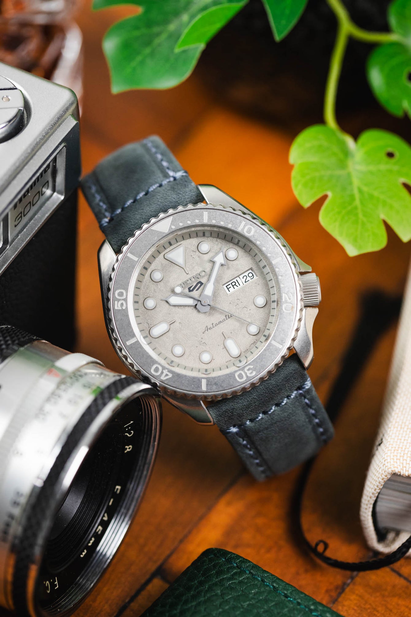 Close-up of a Seiko-style dive watch with a textured light dial and rotating bezel, paired with a blue leather strap with contrasting stitching, set beside a vintage camera and greenery on a wooden surface.