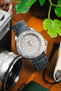 Close-up of a Seiko-style dive watch with a textured light dial and rotating bezel, paired with a blue leather strap with contrasting stitching, set beside a vintage camera and greenery on a wooden surface.