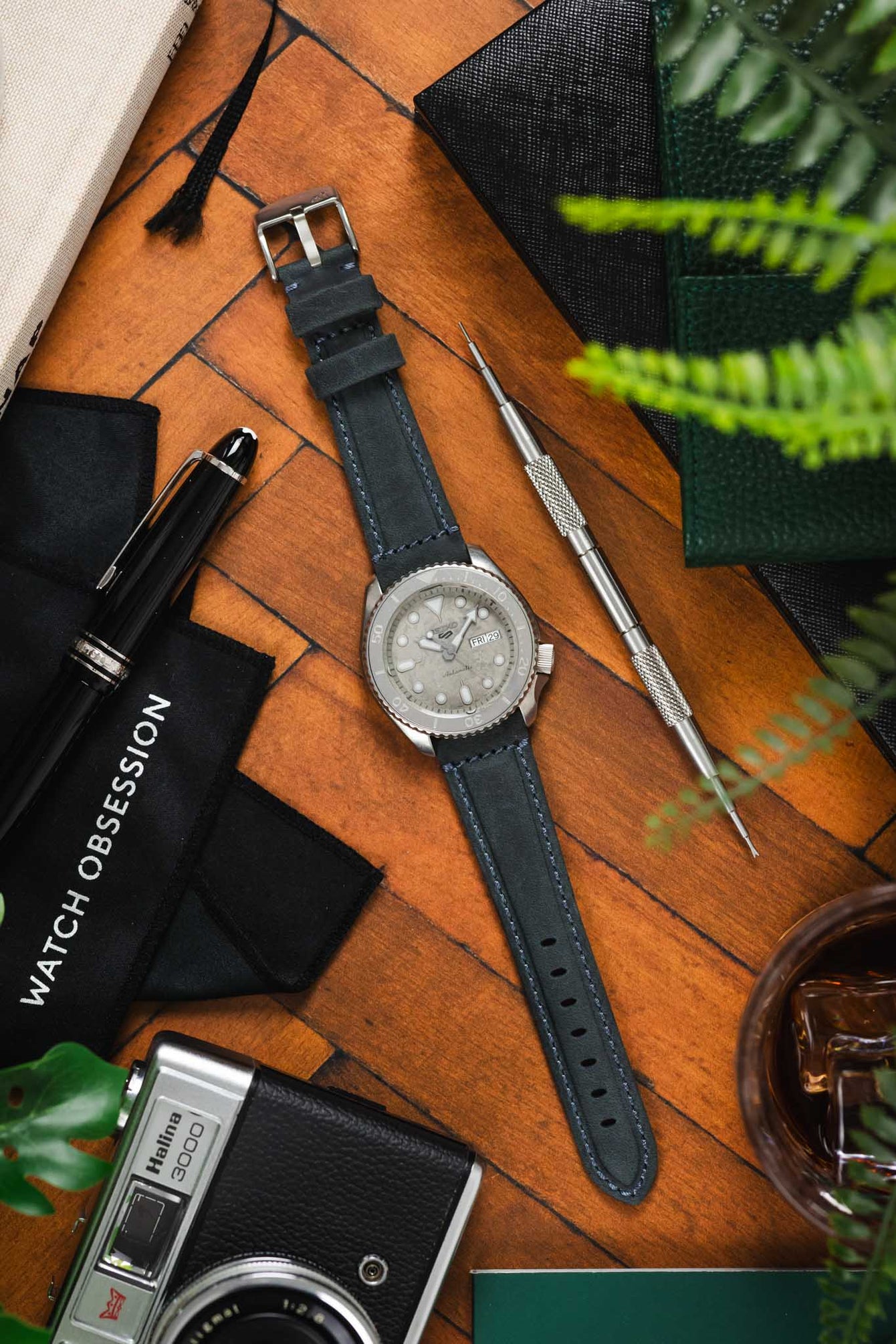 Flat-lay of a Seiko-style dive watch on a blue leather strap with light stitching, surrounded by a camera, tools, pen, greenery, and accessories on a wooden surface.