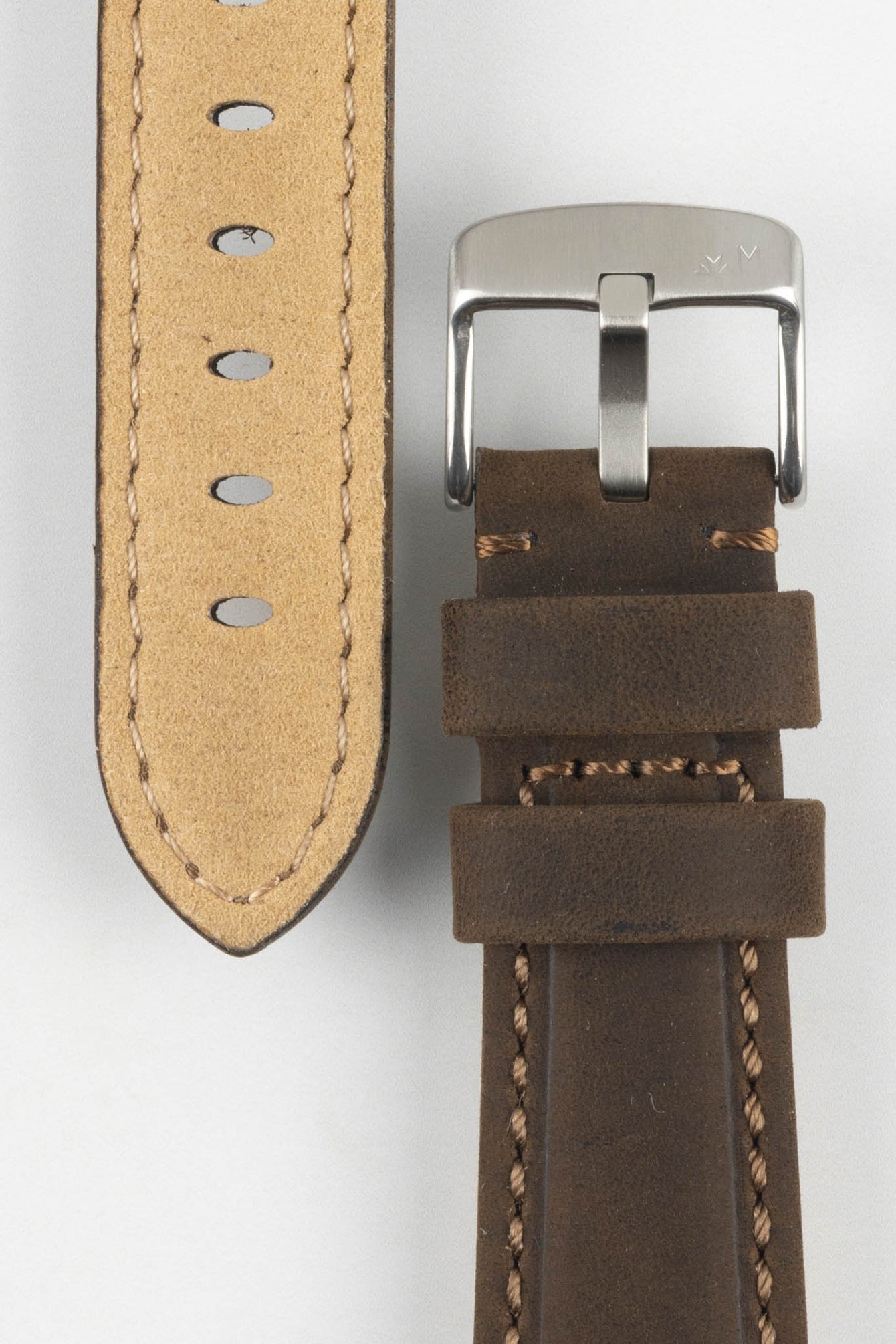 Close-up of a brown leather watch strap with light stitching, showing a silver tang buckle, adjustment holes, and tan suede underside on a light grey background.
