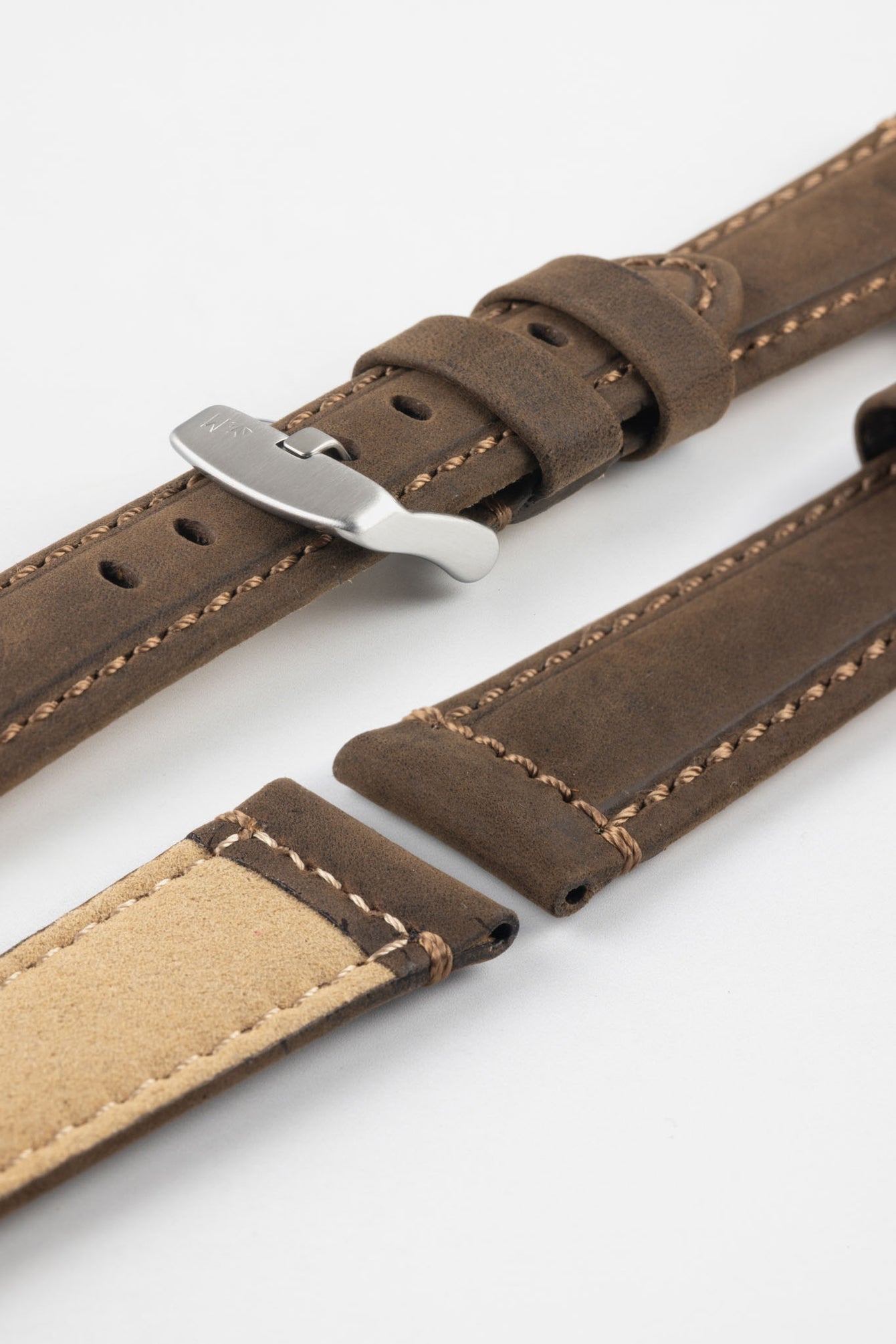 Brown leather watch strap with light stitching, shown in angled close-up with a silver buckle, adjustment holes, and tan suede underside.