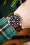 Close-up of a Tudor GMT watch with a black dial and red-and-blue bezel, worn on a wrist with a brown leather strap and green plaid sleeve.