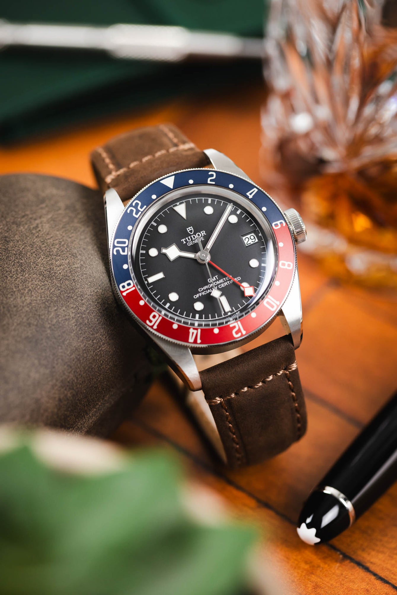 Close-up of a Tudor GMT watch with a black dial and red-and-blue bezel on a brown leather strap, resting on a surface beside a pen and a glass of whisky with a warm, softly blurred background.