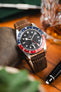 Close-up of a Tudor GMT watch with a black dial and red-and-blue bezel on a brown leather strap, resting on a surface beside a pen and a glass of whisky with a warm, softly blurred background.