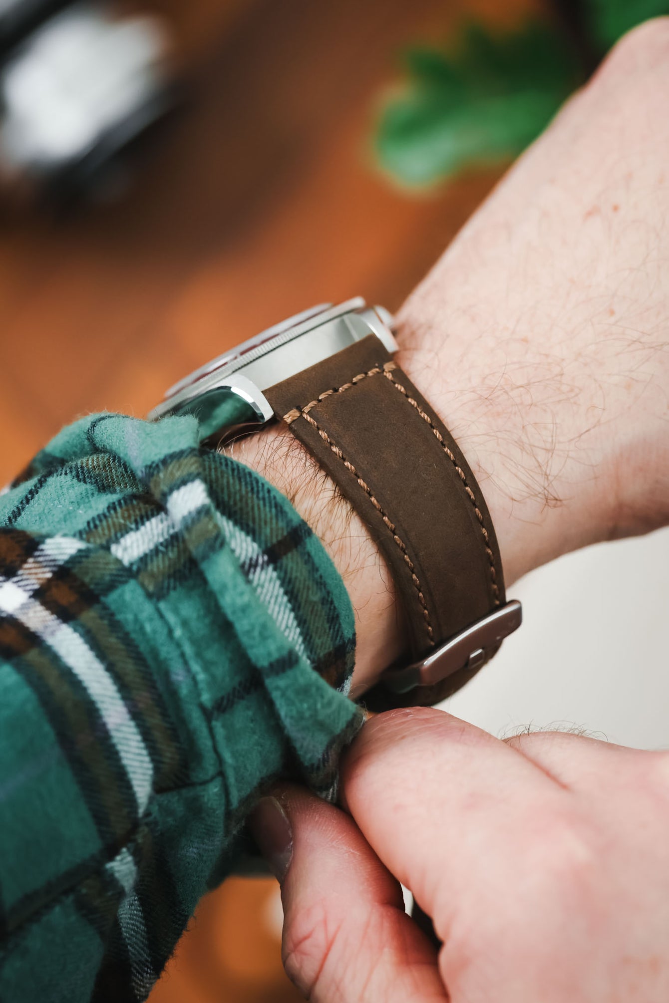 Close-up of a brown leather watch strap with contrast stitching, secured on a wrist, with a hand adjusting the strap and a green plaid sleeve visible.