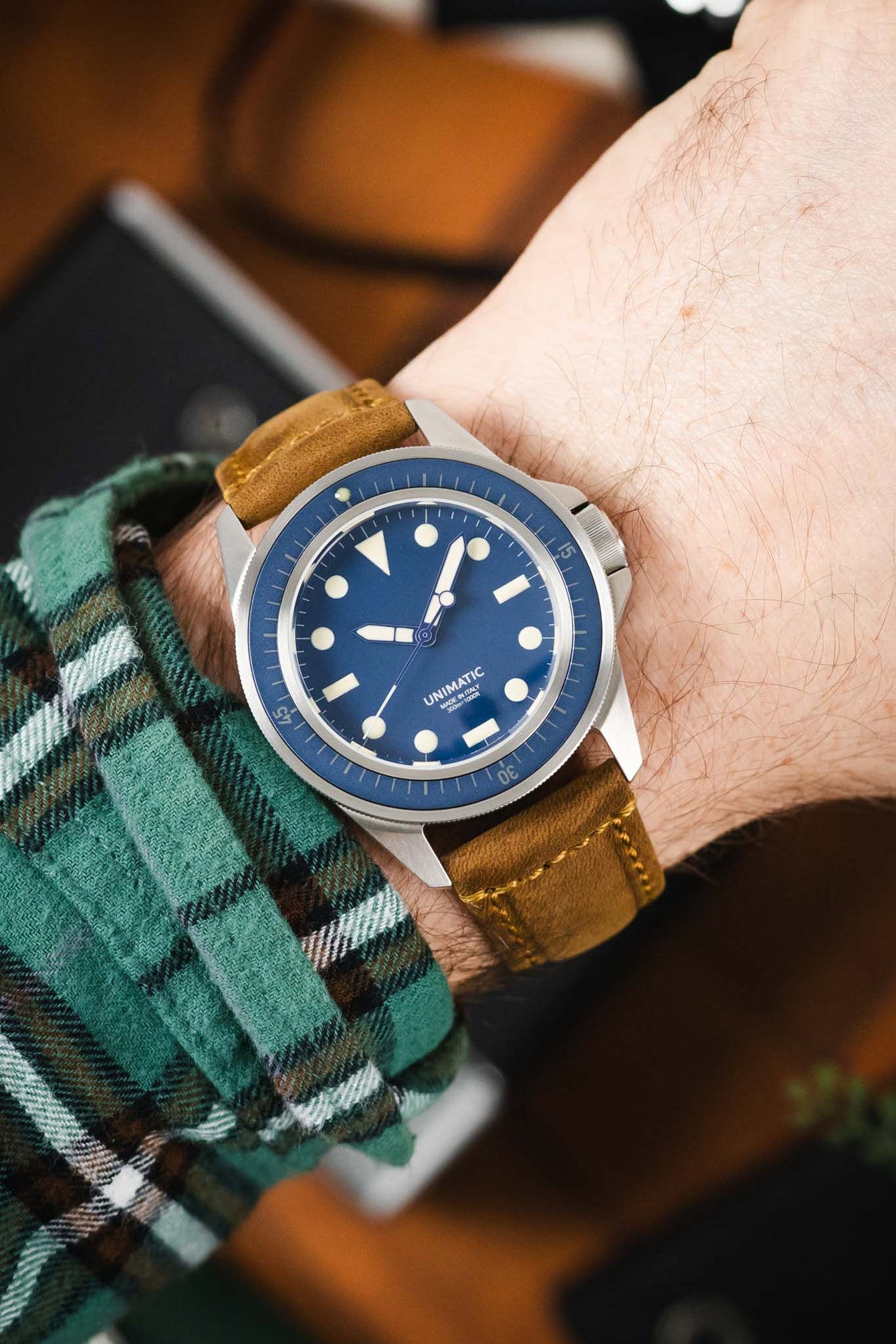 Close-up of a Unimatic dive watch with a blue dial on a tan leather strap, worn on a wrist with a green plaid sleeve.