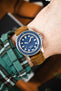 Close-up of a Unimatic dive watch with a blue dial on a tan leather strap, worn on a wrist with a green plaid sleeve.