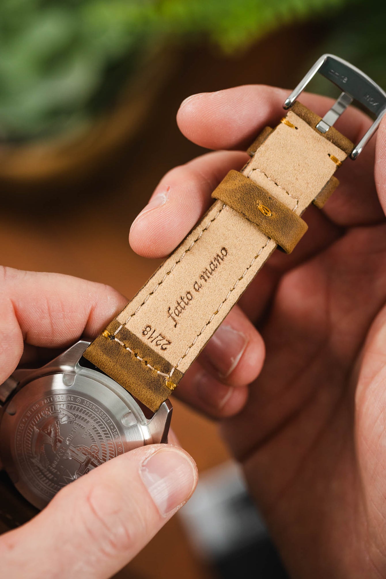 Close-up of the underside of a tan leather watch strap held in hand, showing stitching, keeper loop, and engraved text “Fatto a Mano” and “22/18,” attached to a steel caseback.