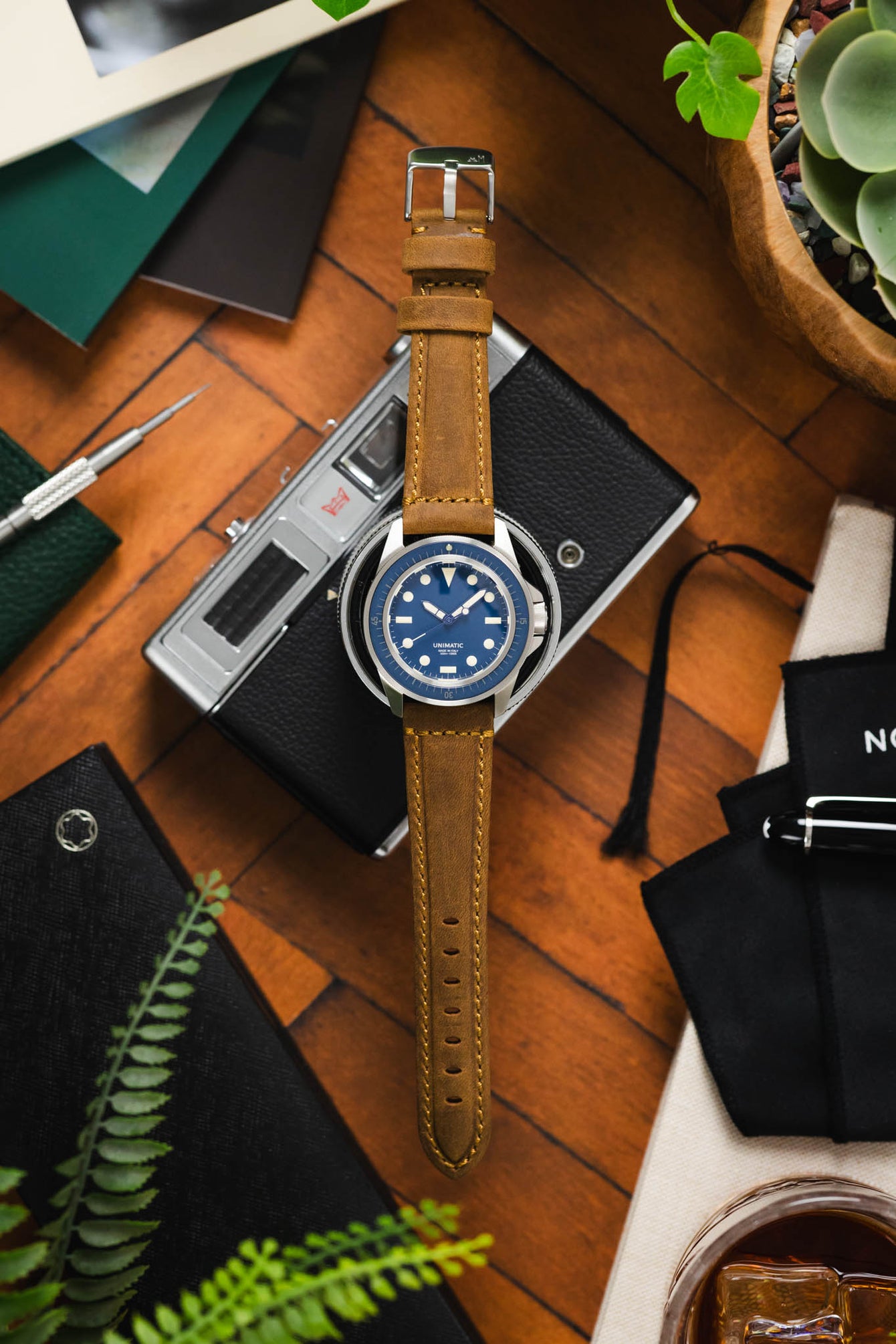 Flat-lay of a Unimatic dive watch with a blue dial on a tan leather strap, placed over a vintage camera and surrounded by tools, greenery, and accessories on a wooden surface.