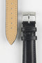 Close-up of a black leather watch strap showing a silver buckle, adjustment holes, and beige suede underside on a light grey background.