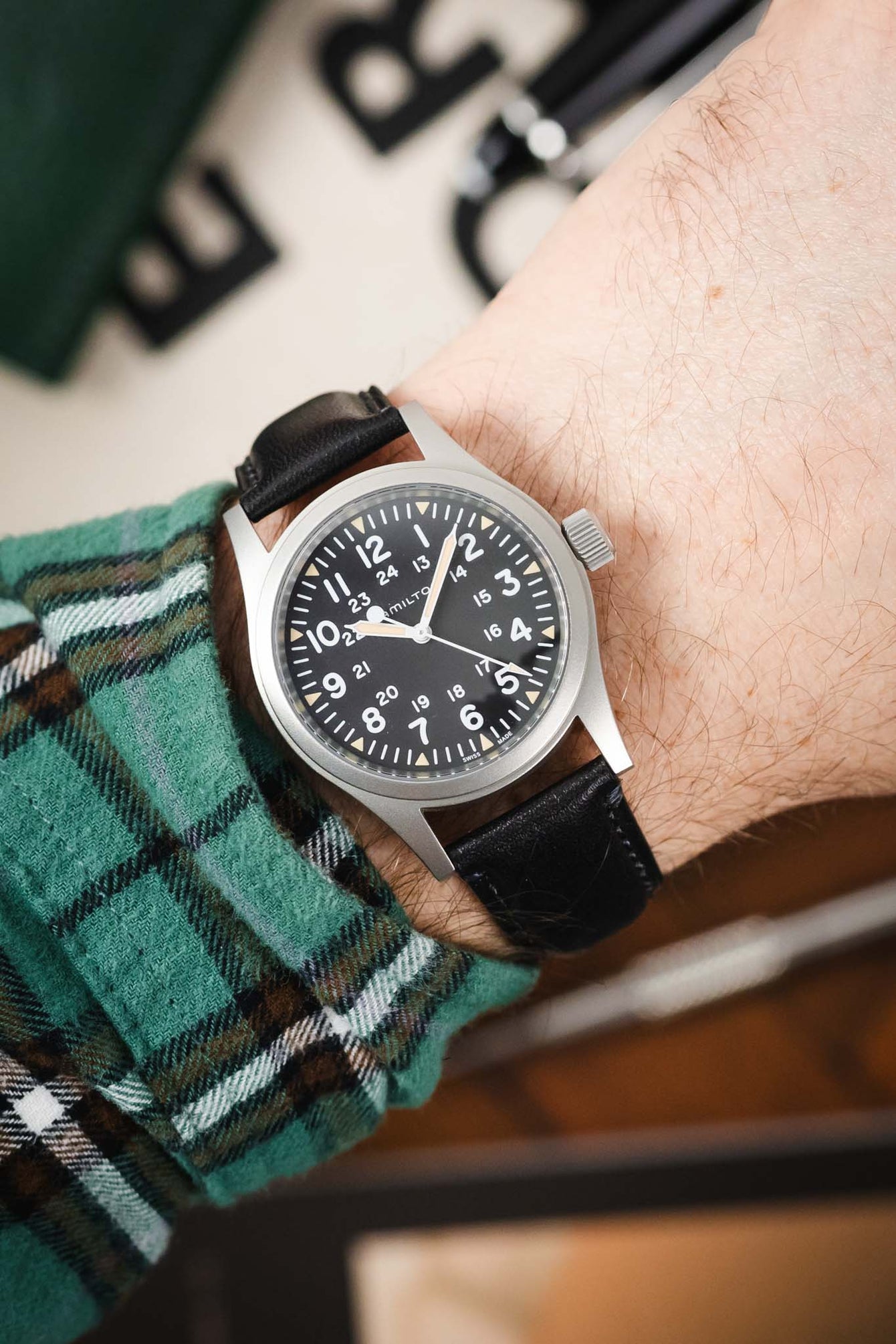 Close-up of a Hamilton field watch with a black dial and white numerals, worn on a wrist with a black leather strap and a green plaid sleeve visible.
