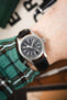 Close-up of a Hamilton field watch with a black dial and white numerals, worn on a wrist with a black leather strap and a green plaid sleeve visible.
